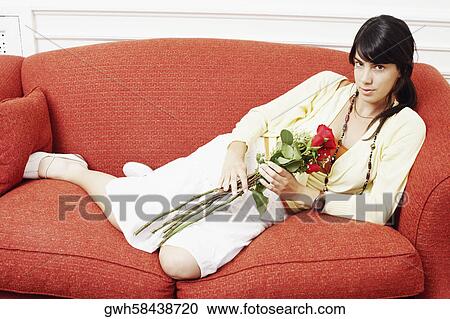 Stock Image - Portrait of a young woman reclining on a couch and holding a bunch of flowers. Fotosearch