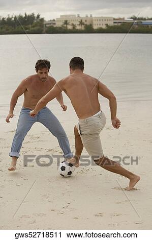 Stock Image - Rear view of a mid adult man playing soccer with a young man on the beach. Fotosearch