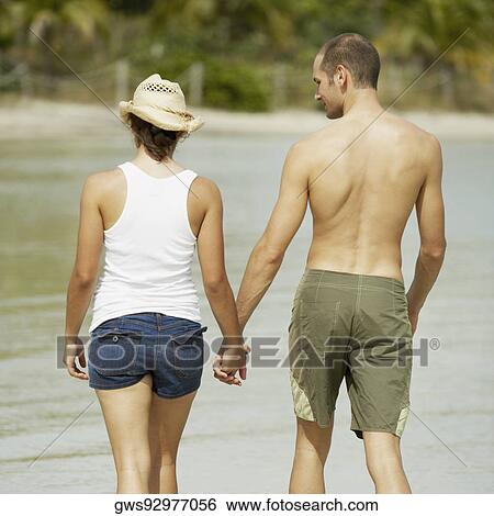 Stock Photograph - Rear view of a young man and a teenage girl holding hands and walking on the beach. Fotosearch