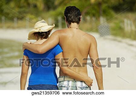Stock Photograph - Rear view of a young man walking on the beach with his arm around a teenage girl. Fotosearch