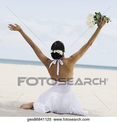 Stock Photography - Rear view of a young woman sitting and holding flowers on the beach. Fotosearch