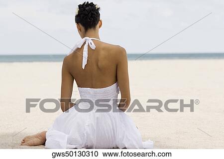 Stock Image - Rear view of a young woman sitting on the beach. Fotosearch