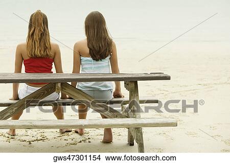 Rear view of two girls sitting on a bench at the beach View Large Photo Image Picture - Rear view of two girls sitting on a bench at the beach. Fotosearch