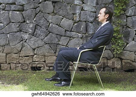 Stock Photograph - Side profile of a businessman sitting on a chair in front of a stone wall. Fotosearch