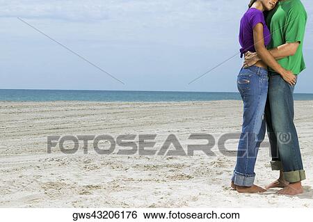 Stock Photograph - Side profile of a young couple embracing each other on the beach. Fotosearch