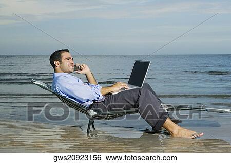 Stock Photograph - Side profile of a young man sitting on a lounge chair on the beach talking on a mobile phone and using a laptop. Fotosearch