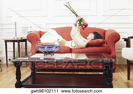 Stock Image - Side profile of a young woman lying on a couch and holding a bunch of flowers. Fotosearch