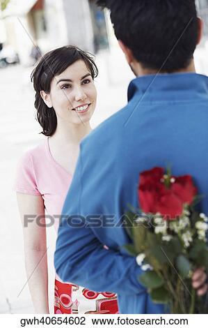 Stock Image - Young man hiding a bunch of roses behind his back with a young woman in front of him. Fotosearch