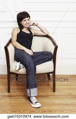 Picture - Young woman sitting in an armchair and talking on a mobile phone. Fotosearch