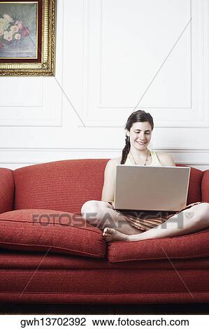 Stock Image - Young woman sitting on a couch and using a laptop. Fotosearch