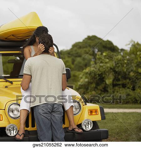 Stock Image - Young woman sitting on the bonnet of a jeep and kissing a mid adult man. Fotosearch