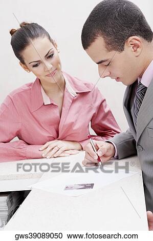 Stock Image - Close-up of a businessman writing on paper with a businesswoman standing beside him. Fotosearch