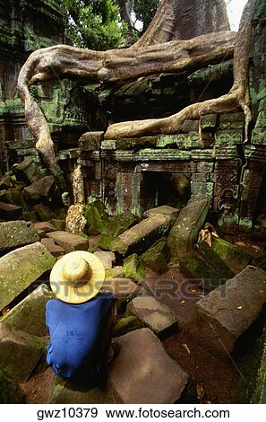 Stock Photo - High angle view of a man crouching on a stone in front of a temple, Ta Prohm Temple, Angkor, Siem Reap, Cambodia. Fotosearch
