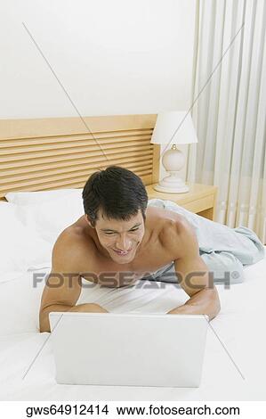 Picture - High angle view of a mid adult man lying on the bed using a laptop. Fotosearch