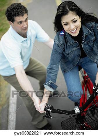 Stock Image - High angle view of a mid adult man helping a mid adult woman ride a bicycle. Fotosearch