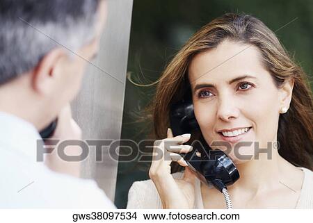Picture - Mid adult woman talking on a pay phone with a mid adult man in front of her. Fotosearch