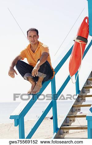 Stock Image - Portrait of a mid adult man sitting on the hand rail of a lifeguard hut and smiling. Fotosearch