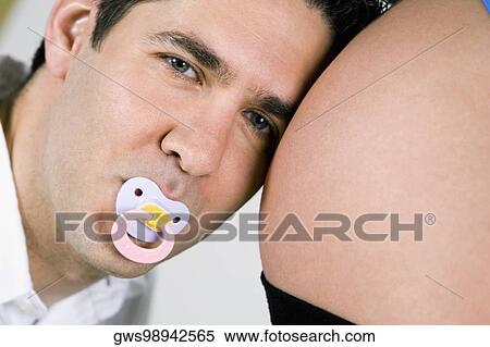 Stock Photography - Portrait of a mid adult man sucking a pacifier and listening to a pregnant woman's abdomen. Fotosearch