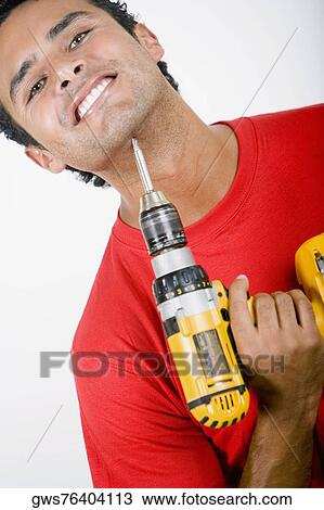 Stock Image - Portrait of a young man holding a drill machine under his chin. Fotosearch