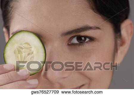 Stock Photo - Portrait of a young woman covering her eye with a cucumber slice. Fotosearch