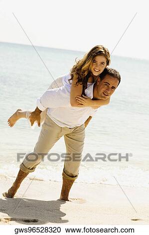 Stock Image - Portrait of a young woman riding piggyback on a mid adult man on the beach. Fotosearch