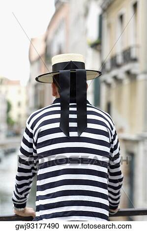 Stock Image - Rear view of a gondolier standing leaning against a railing, Venice, Veneto, Italy. Fotosearch