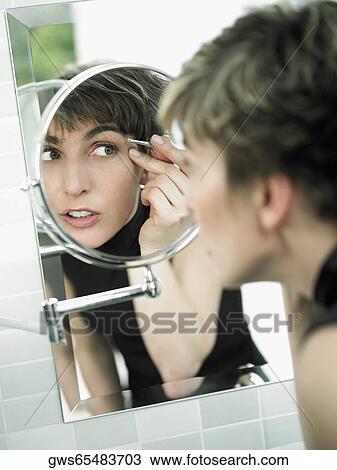 Stock Image - Reflection of a mature woman tweezing her eyebrows in a mirror. Fotosearch