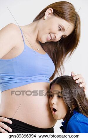 Picture - Side profile of a girl listening to a pregnant young woman's abdomen. Fotosearch