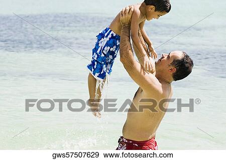 Stock Photo - Side profile of a mid adult man picking up his son in water. Fotosearch