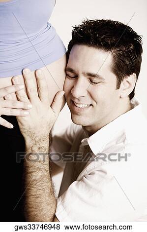 Stock Photo - Side profile of a mid adult man listening to a pregnant young woman's abdomen and smiling. Fotosearch