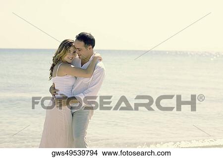 Picture - Side profile of a young woman and a mid adult man embracing each other on the beach. Fotosearch