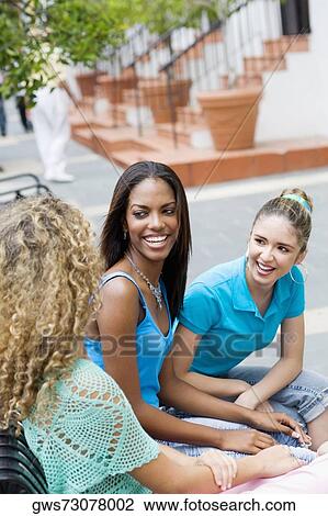 Stock Image - Three teenage girls sitting on a bench and smiling. Fotosearch