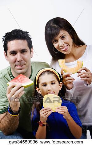 Young woman and a mid adult man with their daughter holding slices of fruits and sticking their tongues out View Large Photo Image Stock Photograph - Young woman and a mid adult man with their daughter holding slices of fruits and sticking their tongues out. Fotosearch