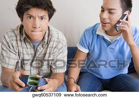 Close-up of a young man playing video game with another young man talking on a mobile phone beside him View Large Photo Image Stock Photo - Close-up of a young man playing video game with another young man talking on a mobile phone beside him. Fotosearch