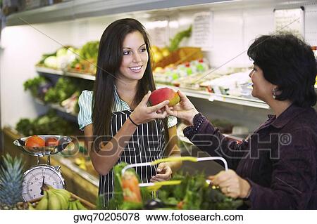 Stock Photo - Female sales clerk giving a mango to a mature woman in a grocery store. Fotosearch