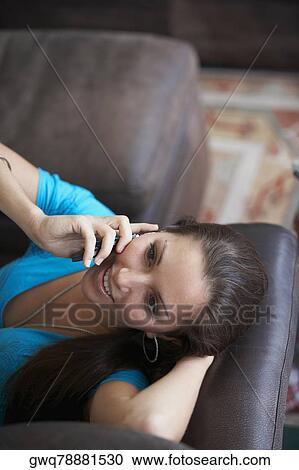 Stock Image - High angle view of a young woman lying on a couch and talking on a mobile phone. Fotosearch