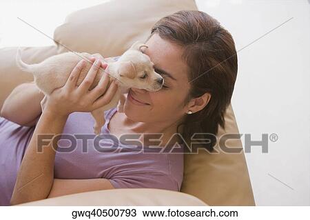 Stock Image - High angle view of a young woman playing with her puppy and lying on a couch. Fotosearch