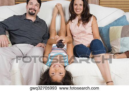 Stock Image - Mid adult couple sitting on a couch with their daughter lying on a couch between them and smiling. Fotosearch