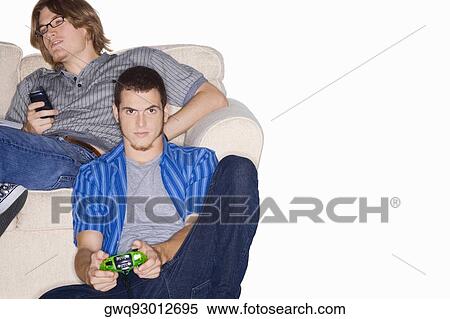 Portrait of a young man playing video game with a remote control and another young man using a mobile phone behind him View Large Photo Image Stock Photography - Portrait of a young man playing video game with a remote control and another young man using a mobile phone behind him. Fotosearch