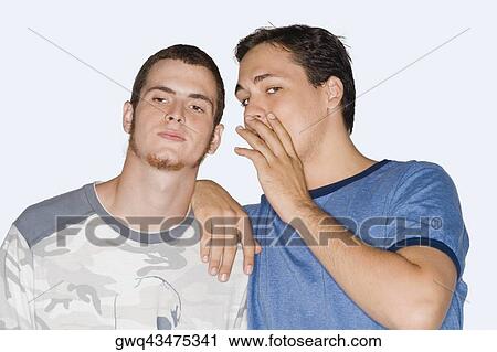 Stock Image - Portrait of a young man whispering into a young man's ear. Fotosearch