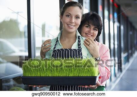 Stock Image - Portrait of a young woman and her mother standing with a tray of wheatgrass and smiling. Fotosearch