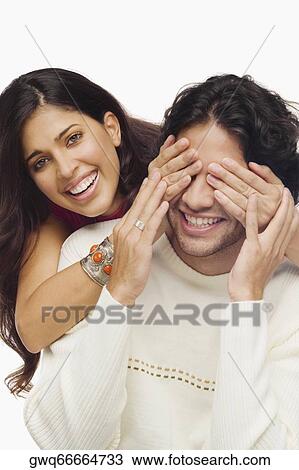 Stock Image - Portrait of a young woman covering eyes of a young man from behind and smiling. Fotosearch