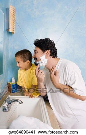 Stock Image - Side profile of a mid adult man shaving with his son sitting beside him in the bathroom. Fotosearch