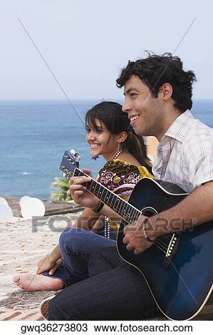Side profile of a young man sitting with a young woman and playing a guitar View Large Photo Image Stock Image - Side profile of a young man sitting with a young woman and playing a guitar. Fotosearch