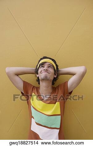 Picture - Young man leaning against a wall with his hands behind his head. Fotosearch