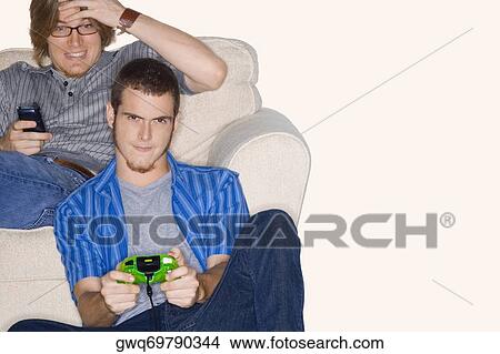 Picture - Young man playing video game with another young man using a mobile phone behind him. Fotosearch