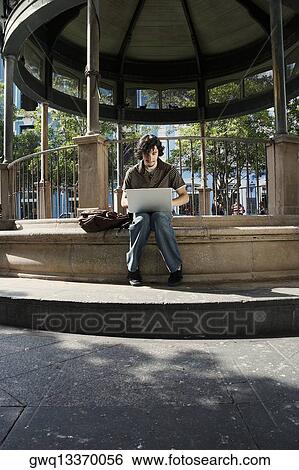 Young man sitting and using a laptop View Large Photo Image Stock Photograph - Young man sitting and using a laptop. Fotosearch