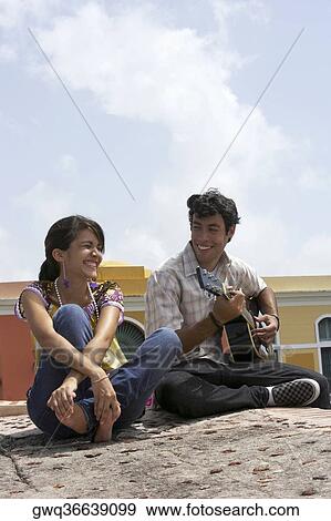 Stock Photo - Young man sitting with a young woman and playing a guitar. Fotosearch