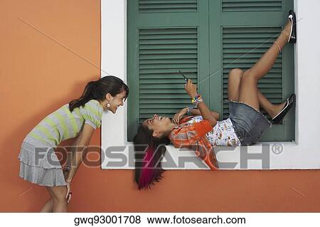 Stock Photo - Young woman lying on a window sill and text messaging with another young woman looking at her. Fotosearch