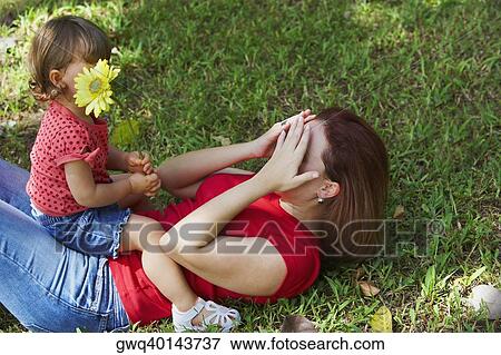 Young woman lying on grass and covering her face with her hands and her daughter sitting on her stomach View Large Photo Image Stock Photo - Young woman lying on grass and covering her face with her hands and her daughter sitting on her stomach. Fotosearch